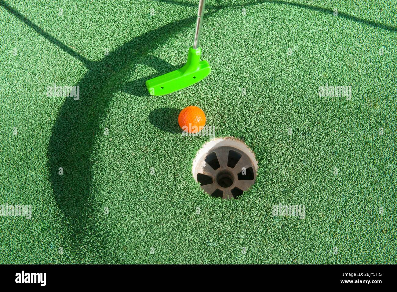 Green mini golf club puts and orange ball towards a hole on green turf Stock Photo