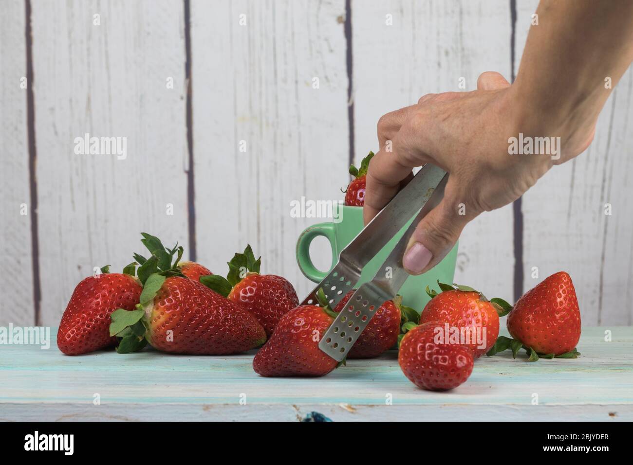 Human hand picking strawberries with pliers, on a white wooden background Stock Photo