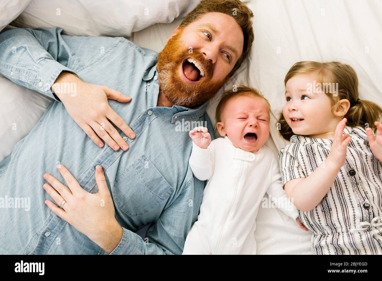 Laughing father lying on bed with crying baby boy (2-3 months) and daughter (2-3) Stock Photo