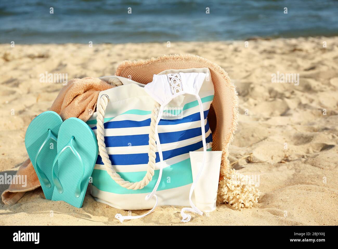 Bag with flip-flops, swimsuit, hat, towel and cosmetics on sand beach ...