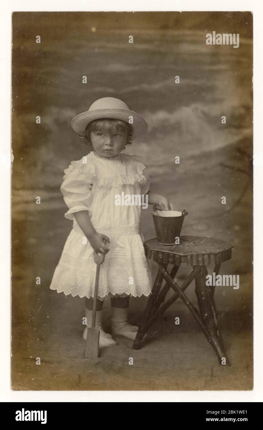 Early 1900's seaside postcard of young child boy dressed as a girl as was normal for the time.) holding a bucket and spade, Eastbourne, Sussex, England U.K. circa 1907 Stock Photo
