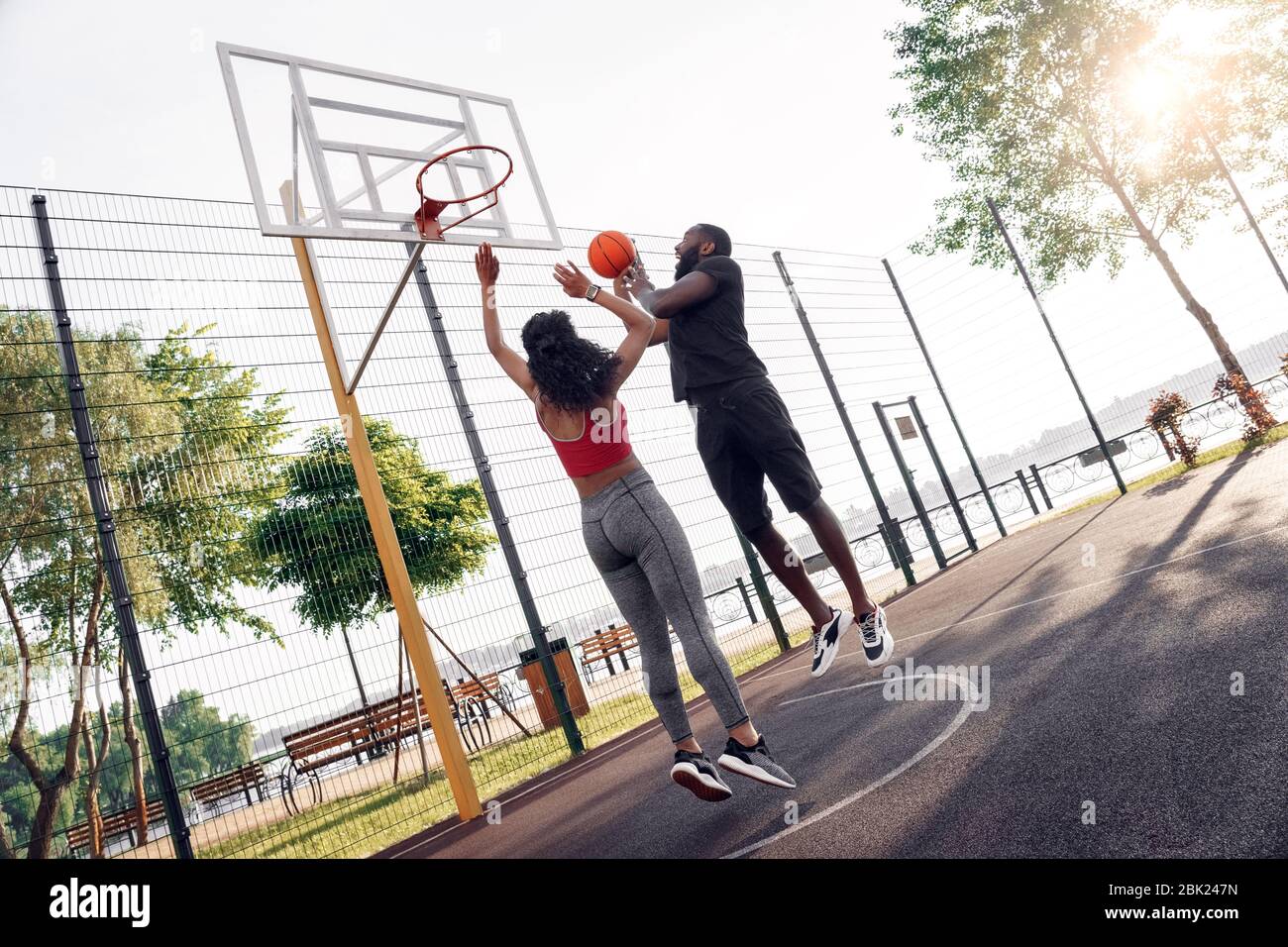 Outdoors Activity. African couple on basketball court guy throwing ball to  basket excited while girl jumping blocking Stock Photo - Alamy