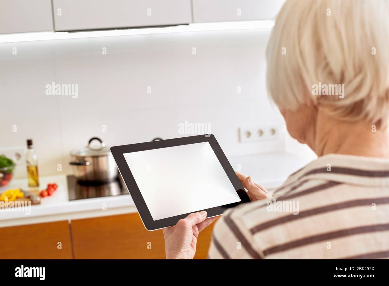 Pensioner woman using modern tablet computer and standing in kitchen Stock Photo