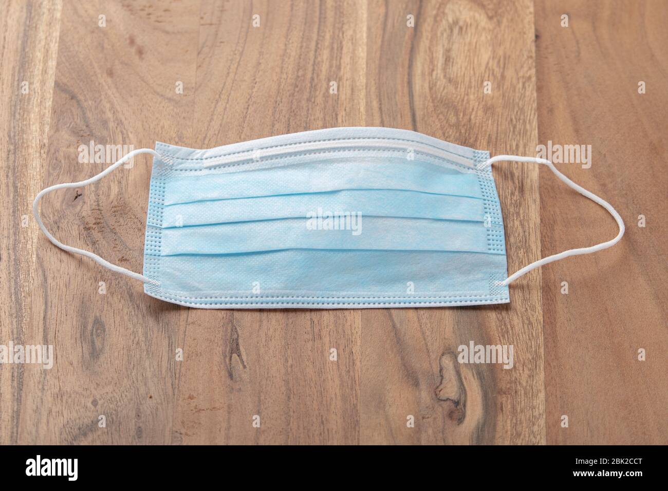 Photo of a blue surgical mask placed on a wooden table made of solid brown oak. The mask with its blue side facing upwards and is ready to be used. Stock Photo