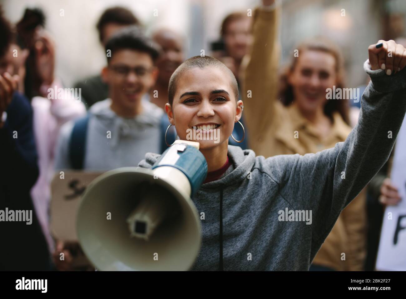 Young woman standing outdoors with group of demonstrator in background. Woman protesting with a megaphone outdoors on road. Stock Photo