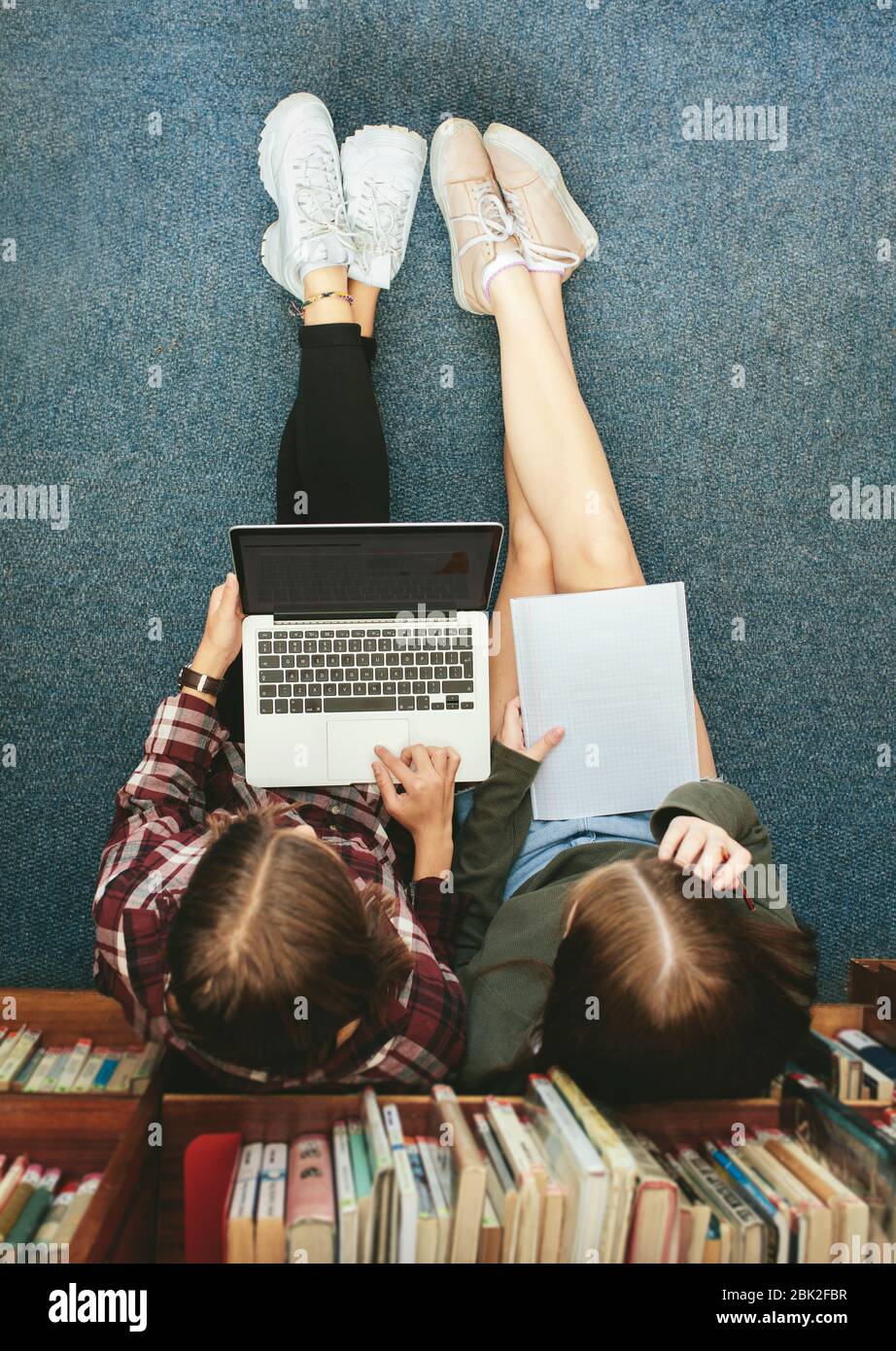 Top view of two female students sitting on floor by a bookshelf and working on a laptop. Two girl school students studying together in library. Stock Photo
