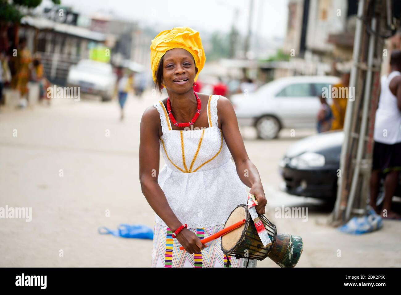 abidjan, ivory coast - February 13, 2018: Young Ivorian woman in white camisole, her head tied with a piece of yellow loincloth, taps a drum that she Stock Photo