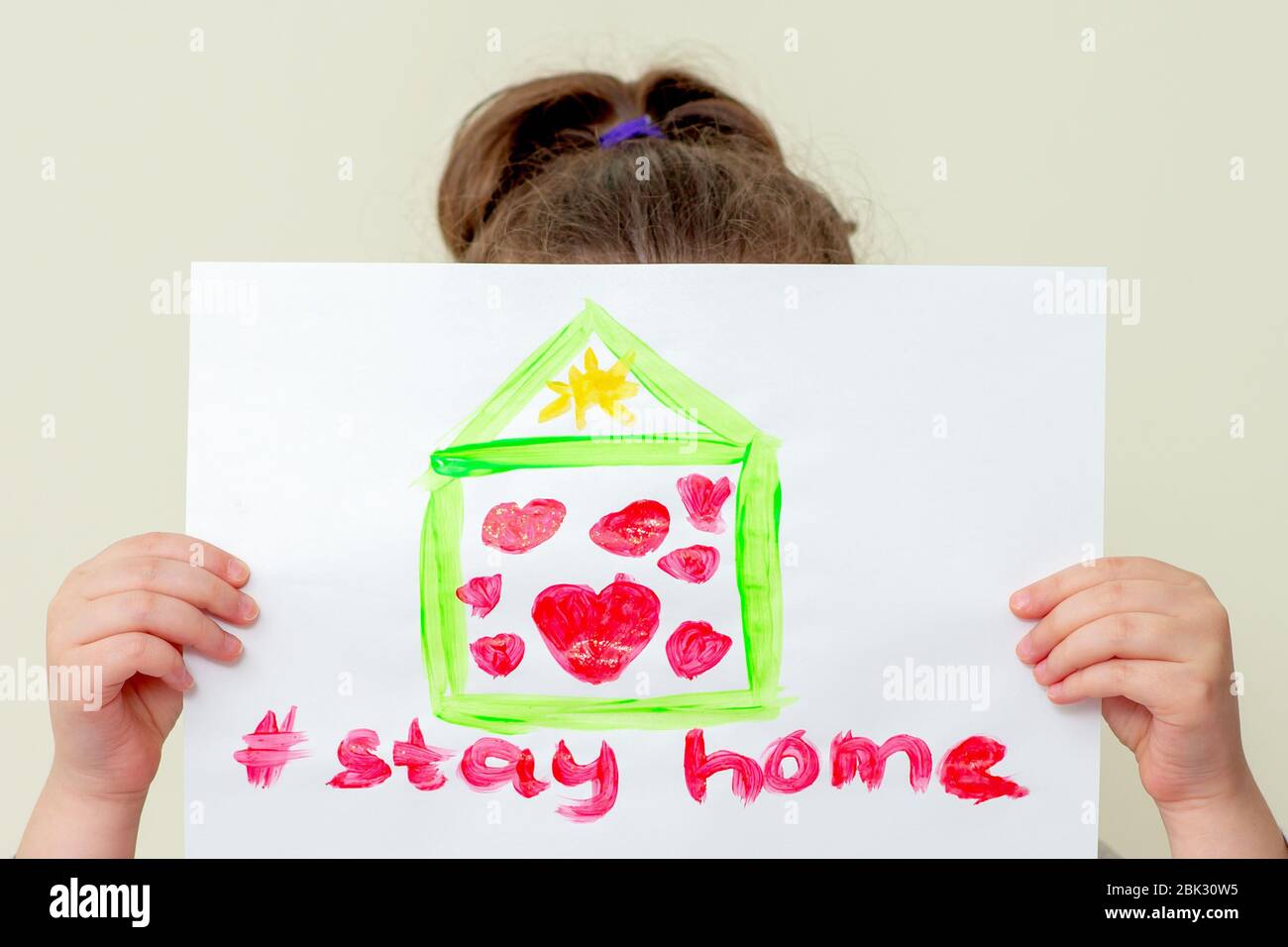 Little girl covers his face a picture of a house with hearts and words Stay Home on sheet of paper, close-up. Stay home concept. Stock Photo