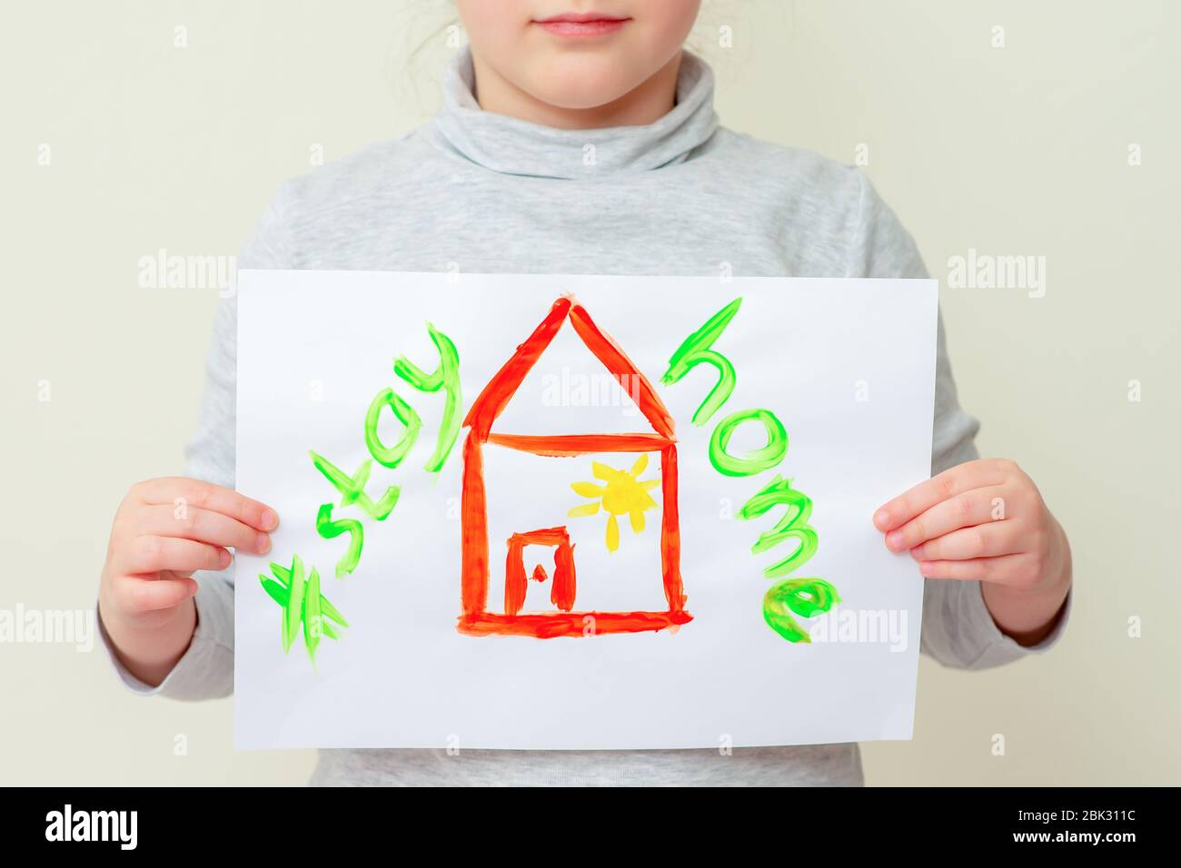 Little girl holds picture red house with words Stay Home on sheet of paper, close-up. Stock Photo