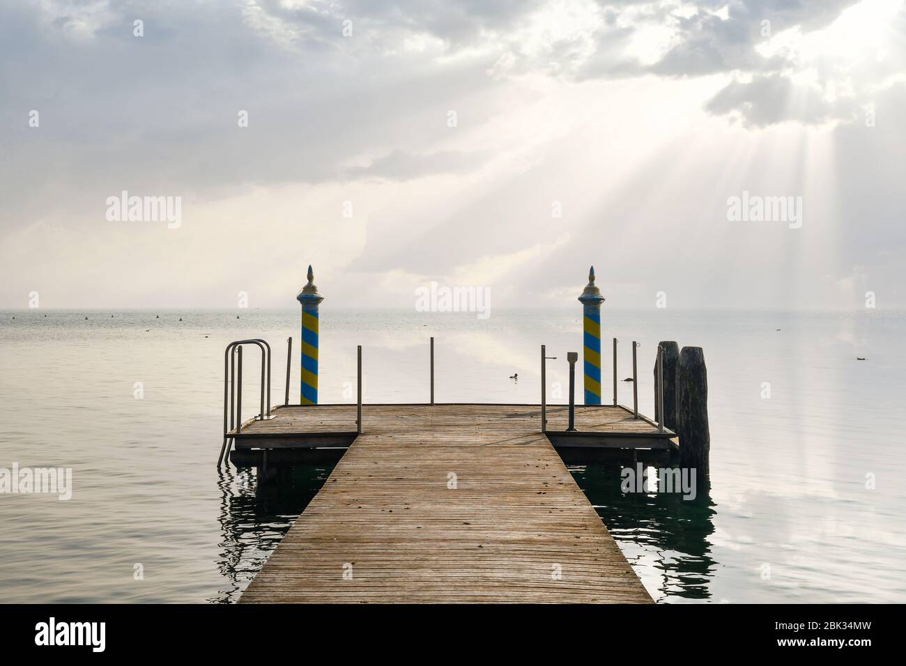 A wooden pier on the lake shore with a dramatic sky and sun rays streaming through the clouds and reflected on the water, Lake Garda, Verona, Italy Stock Photo