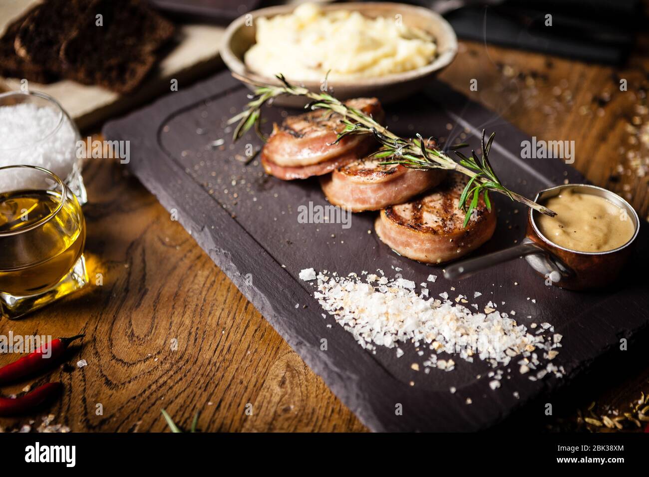 Pork tenderloin with mashed potatos, bacon and mustard sauce covered with a branch of smoking rosemary on black board. Delicious healthy traditional Stock Photo