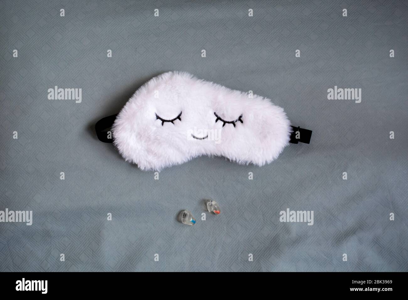 White cute sleep mask made of fluffy faux fur in a cloud shape with closed eyes embroidered on it and custom molded sleep ear plugs on a blue grey pil Stock Photo