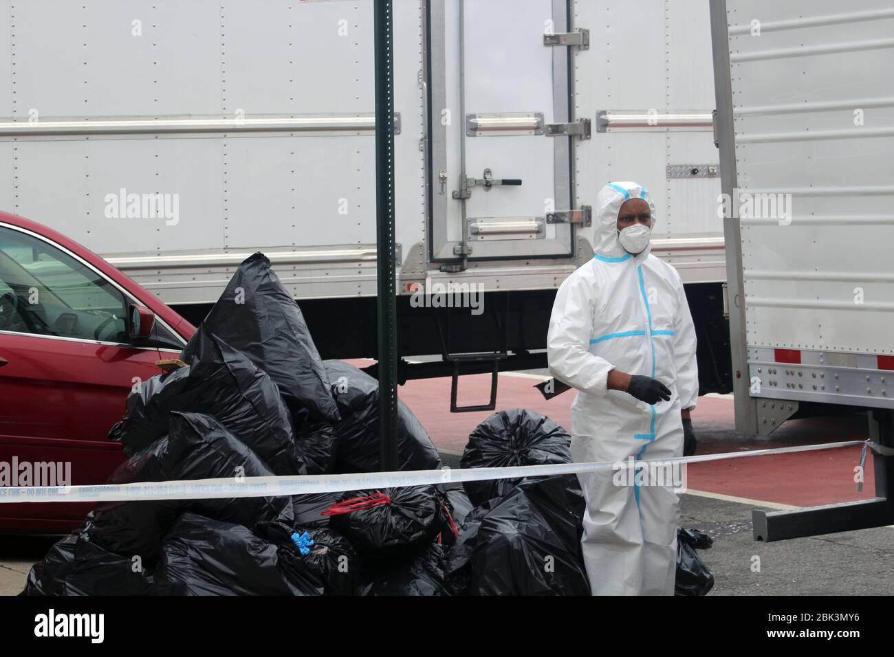 April 30, 2020, New York, New York, USA: This man, dressed in a hazmat, stands in the street surrounded by trucks. Behind him, big black garbage bags have been piled up in front of the Andrew T. Cleckley Funeral Home in Brooklyn where bodies non properly cared are removed. 60 corpses were stored in 4 non-refregerated rented trucks lining the street nearby stores. Neighbors reported a foul smell still persistent and fluids dripping from the trucks. Some remains were also found lying on the facilityÃ¢â‚¬â„¢s floor. The funeral home was overwhelmed by COVID-19 deaths, waiting for weeks for cremat Stock Photo