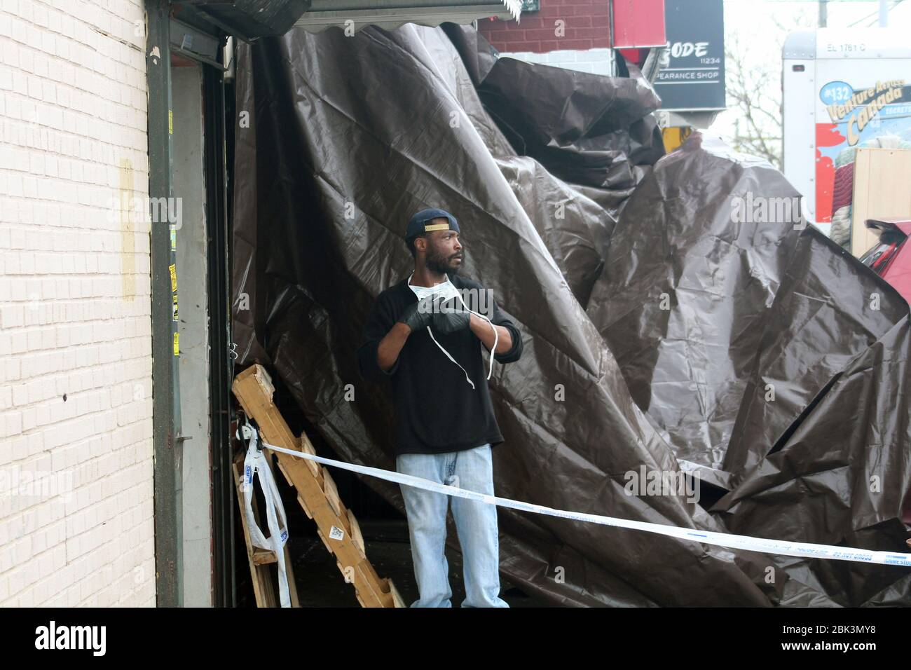 April 30, 2020, New York, New York, USA: A worker, mask face down, looks towards the street along a brown tarp set up at the entrance of the Andrew T. Cleckley Funeral Home in Brooklyn where bodies non properly cared are removed. 60 corpses were stored in 4 non-refregerated vehiciles lining the street nearby stores. Neighbors reported a foul smell still persistent and fluids dripping from the trucks. The funeral home was overwhelmed by COVID-19 deaths, waiting for weeks for cremations in New York. (Credit Image: © Marie Le Ble/ZUMA Wire) Stock Photo