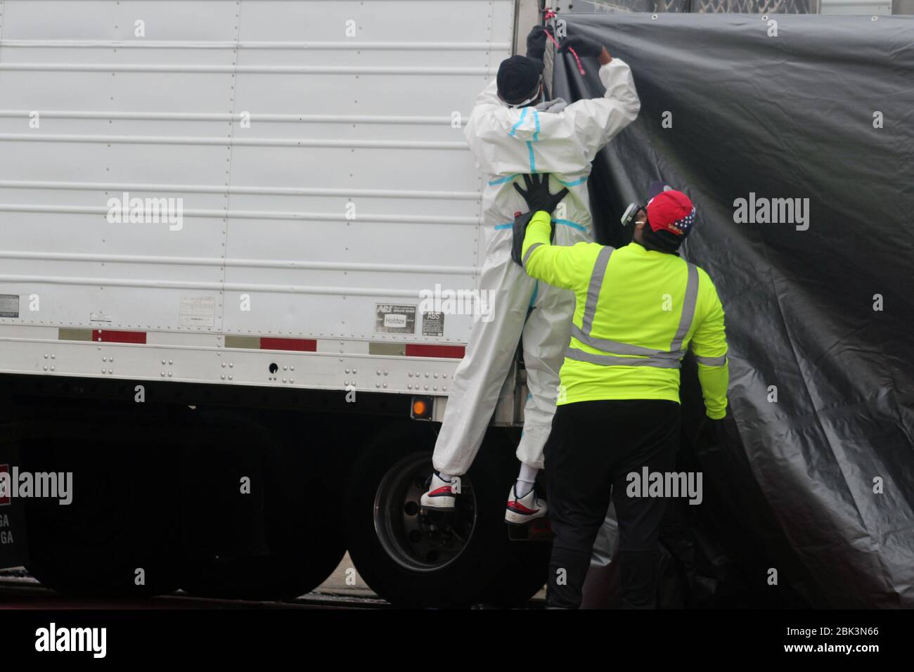 April 30, 2020, New York, New York, USA: The man in a yellow t-shirt supports with his hand his co-worker dressed in a hazmat, who fixes a tarpaulin at the back of a big refrigerated truck to be out of sight next to the Andrew T. Cleckley Funeral Home in Brooklyn where corpses not properly cared have been removed. 60 bodies were stored in 4 non-refregerated vehiciles lining the street nearby stores. Neighbors reported a foul smell still persistent and fluids dripping from the trucks. The funeral home was overwhelmed by COVID-19 deaths, waiting for weeks for cremations in New York. (Credit Imag Stock Photo