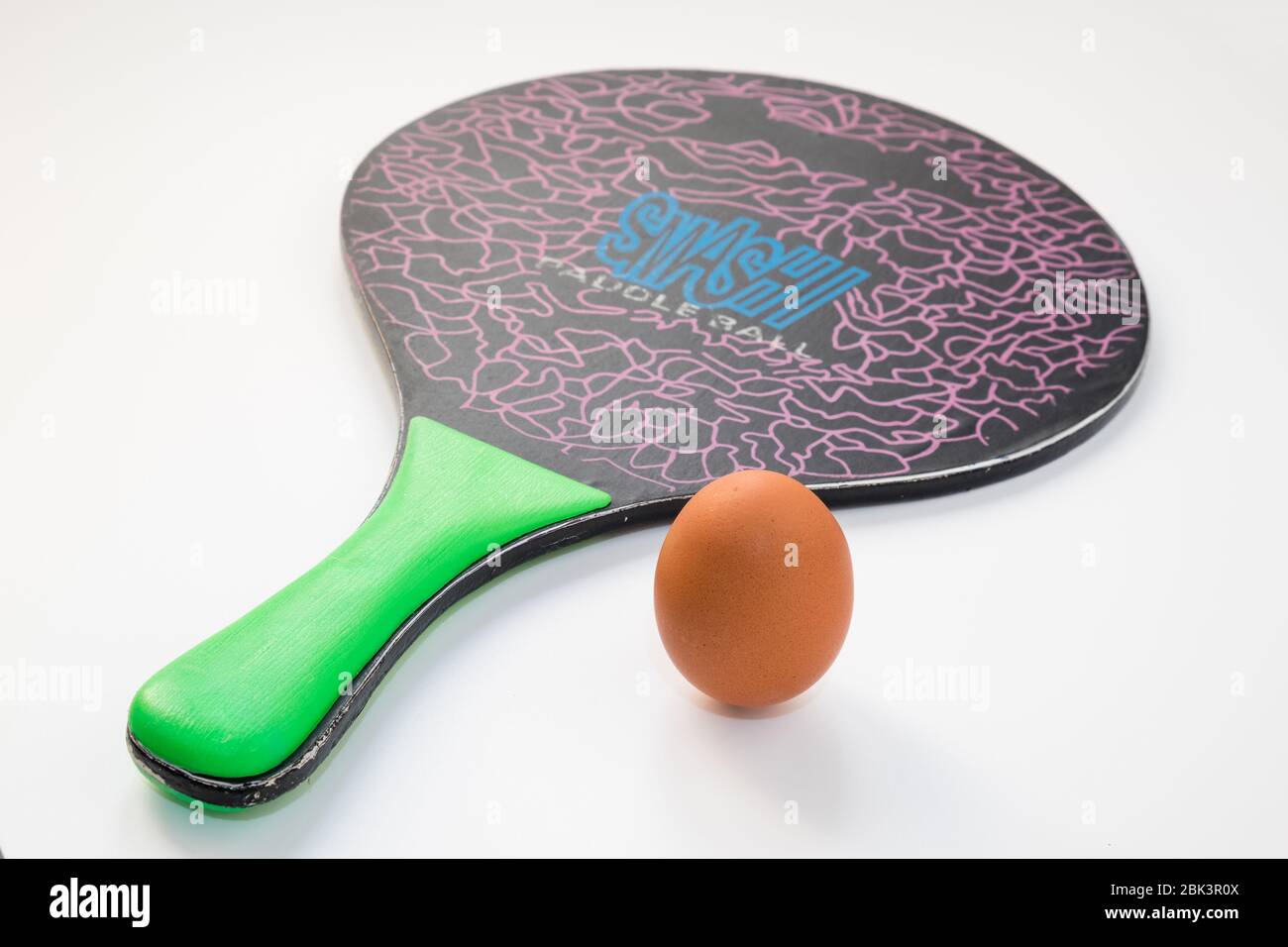 An egg rests on white background near a wooden beach tennis racket, a fragile versus hard concept or sports power supply. Stock Photo