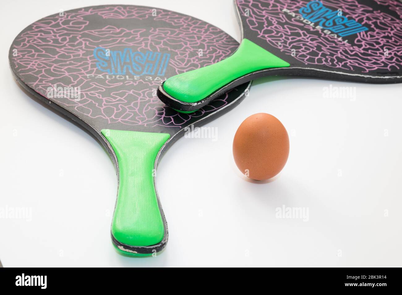 An egg is resting and replaces the ball in the middle between two beach tennis rackets on a white background, the composition expresses the concept of Stock Photo