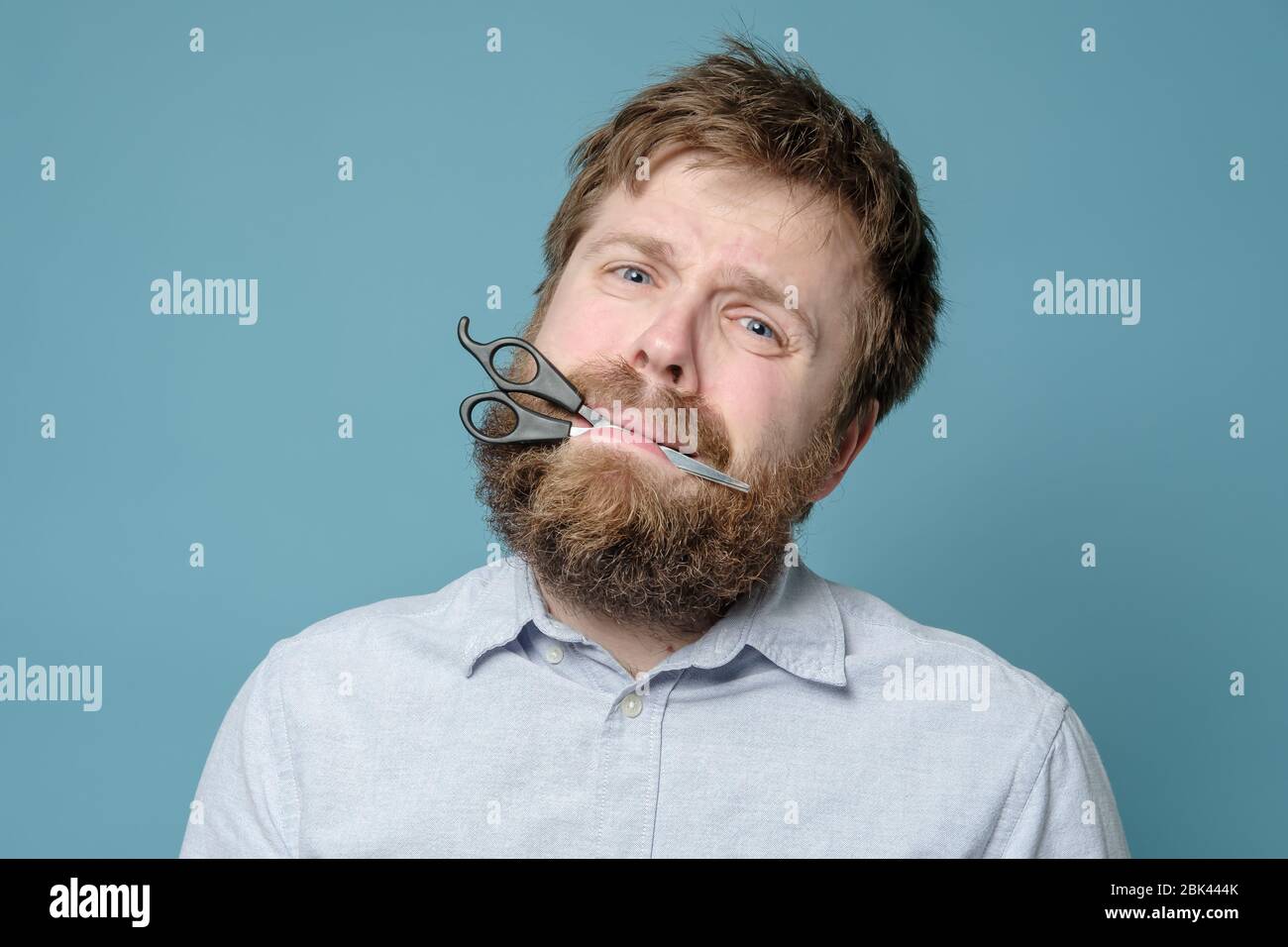 Unhappy shaggy, bearded man with scissors in his mouth in hysteria, he had not been in the barbershop for a long time. Quarantine Stock Photo