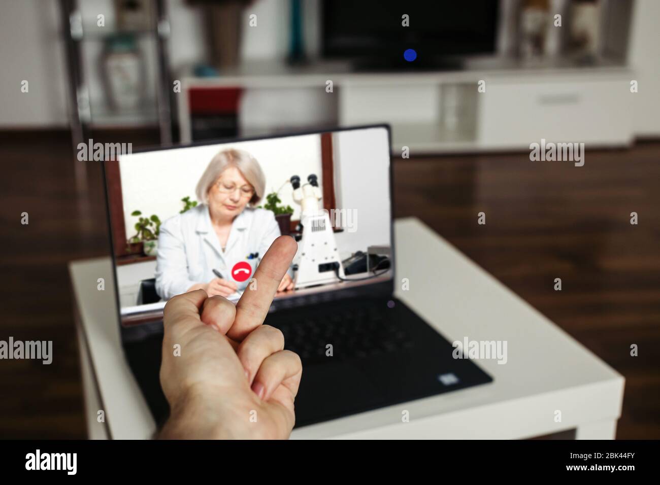 Telemedicine negative concept. Angrey male showing middle finger to a doctor GP on a computer screen, living room home background Stock Photo