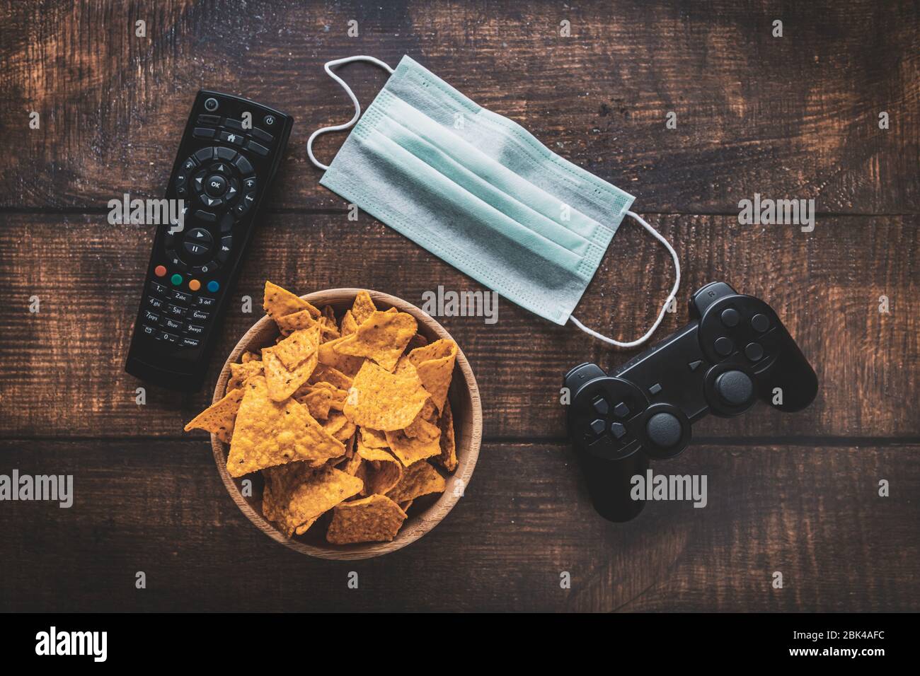 Remote control, game console rc, bowl of crisps and medical mask on wooden table. Coronavirus Covid-19 quarantine stay at home concept. Stock Photo