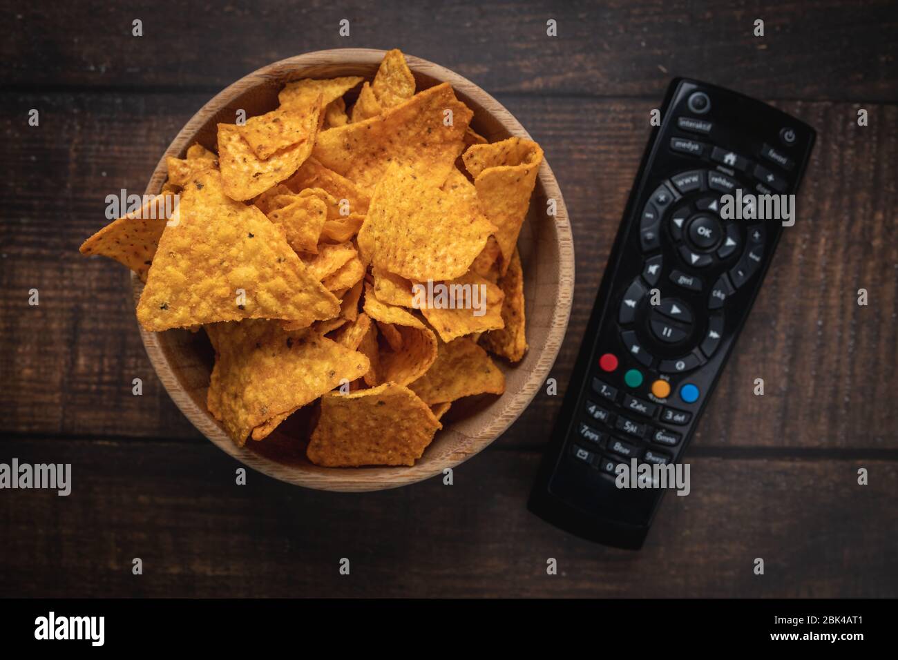 Chips in a wooden bowl next to the TV remote control against a burnt wooden background. Top view. Close up. Toned. Stock Photo