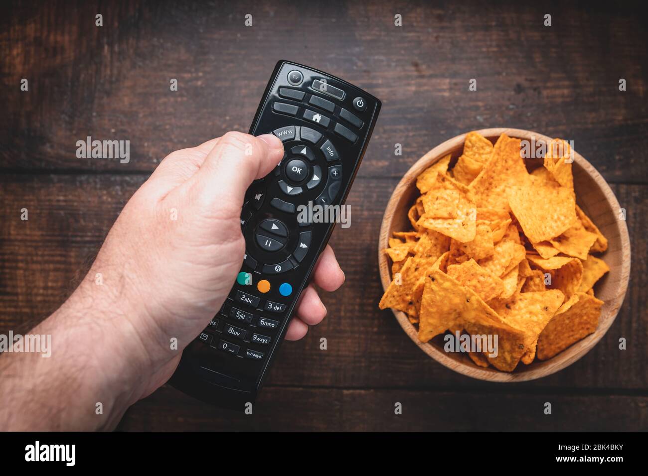 Chips in a wooden bowl next to the TV remote control against a burnt wooden background. Top view. Close up. Toned. Stock Photo