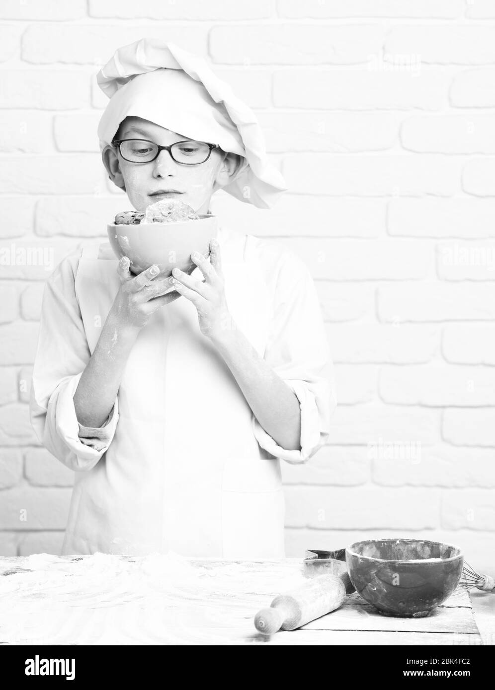 young boy cute cook chef in white uniform and hat on stained face flour with glasses standing near table with rolling pin and holding turquoise bowl with chocolate cookies, on brick wall background Stock Photo
