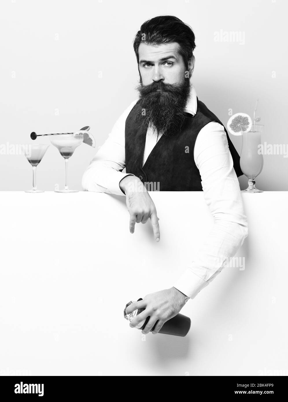 handsome bearded barman with long beard and mustache has stylish hair on serious face holding shaker and made alcoholic cocktail in vintage suede leather waistcoat, on purple green studio background Stock Photo