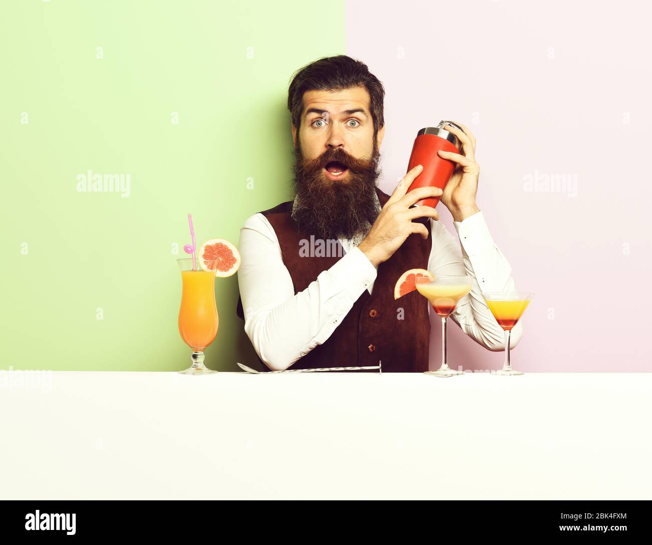 handsome bearded barman with long beard and mustache has stylish hair on surprised face holding shaker and made alcoholic cocktail in vintage suede leather waistcoat, on purple green studio background Stock Photo