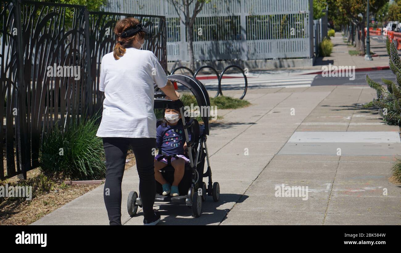 Asian-American mother walking her baby or toddler in a stroller during COVID19 shutdown, both are wearing a mask as they stroll in Emeryville, CA, USA. Stock Photo