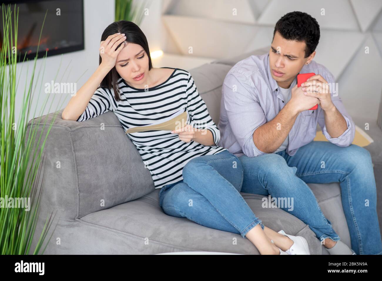Anxious woman with letter, a husband next with smartphone. Stock Photo