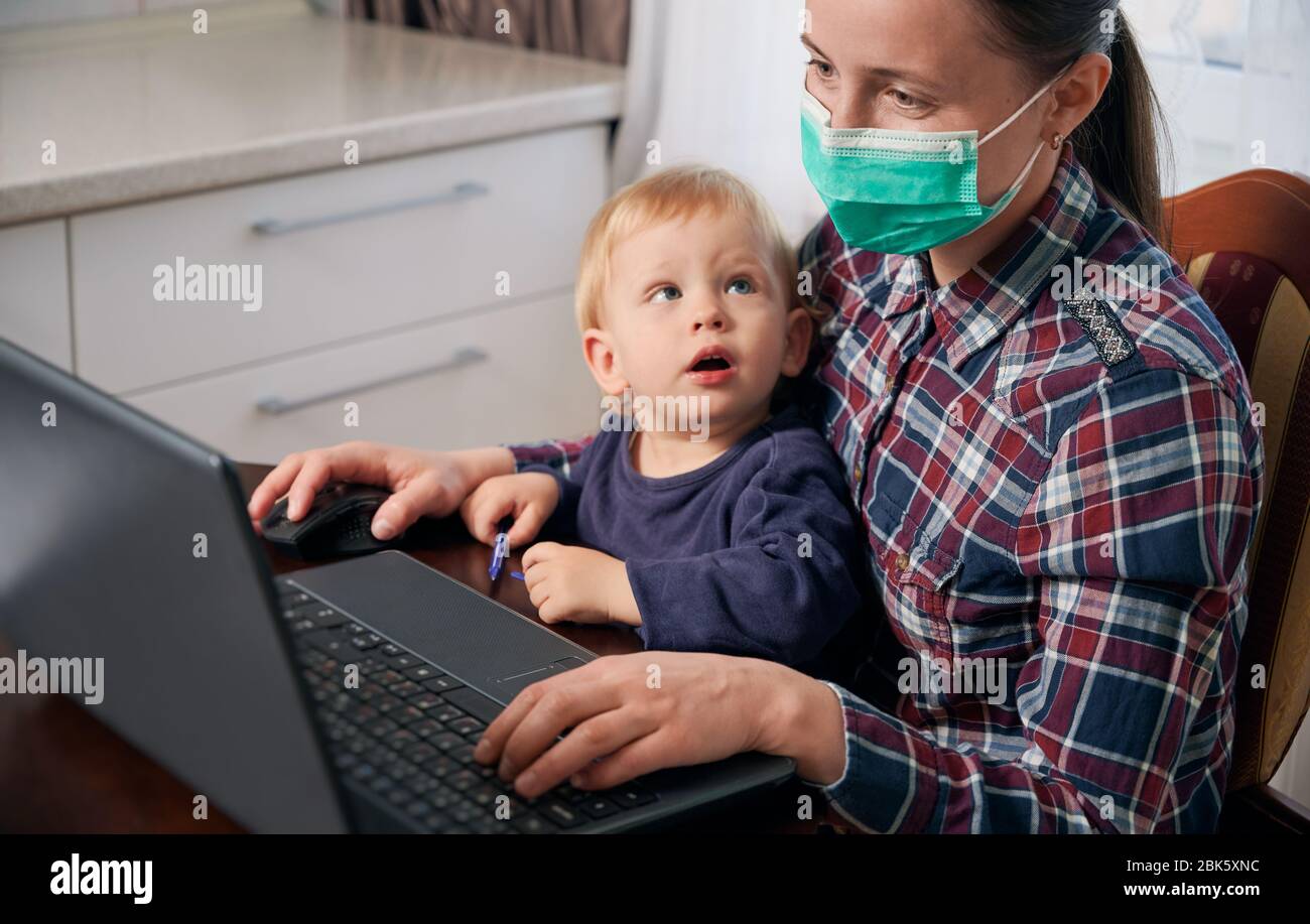 Mother stays at home wearing mask, working remotely on laptop by the table with toddler on her laps. Curious little boy. Home office with kids during global pandemic situation. Covid-19 concept Stock Photo