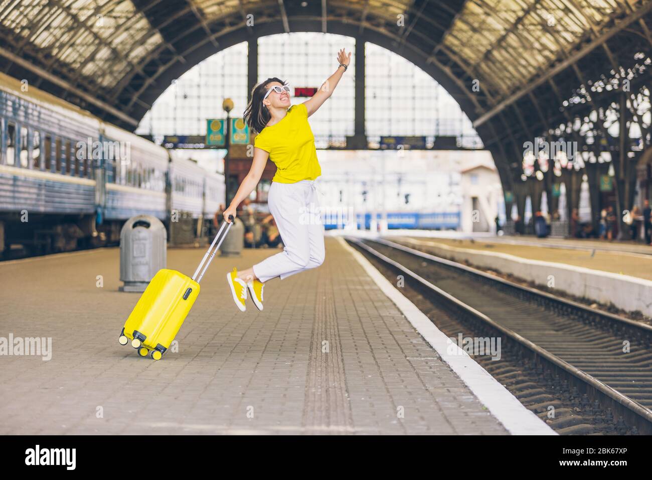 woman jumping at railway station platform with yellow suitcase Stock