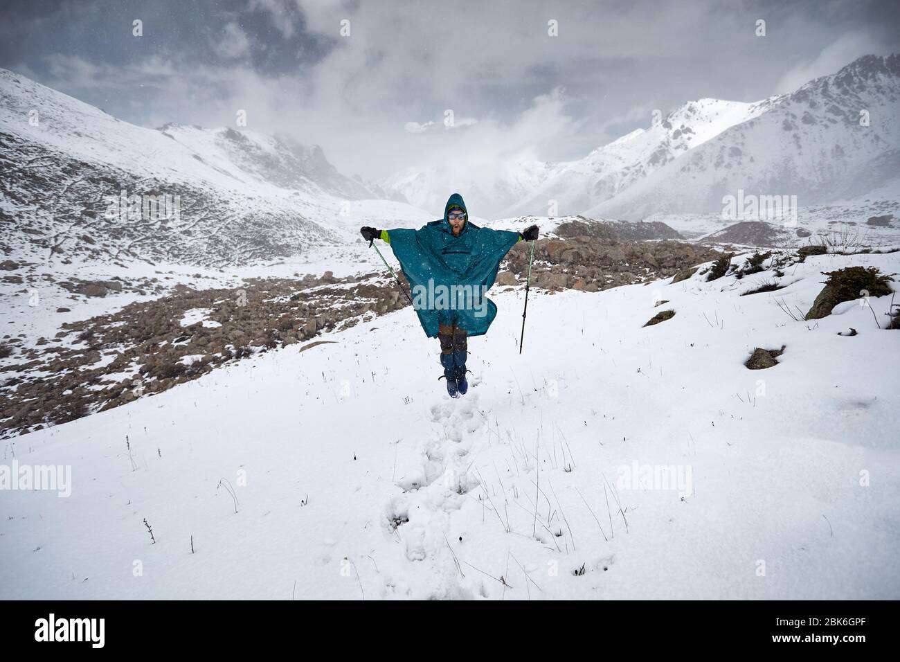 Hiker in blue raincoat poncho is jumping in snowy mountains. Outdoor travel concept Stock Photo