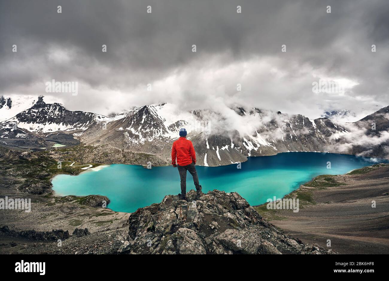 Man in red jacket is looking at Ala-Kul Lake in the Tien Shan mountains with white foggy clouds in Karakol national park, Kyrgyzstan Stock Photo
