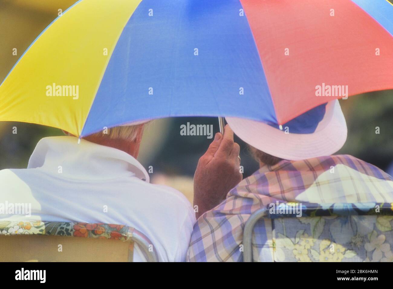 Elderly couple sitting under an umbrella offering protection from the heat and sunshine, England, Britain, UK Stock Photo