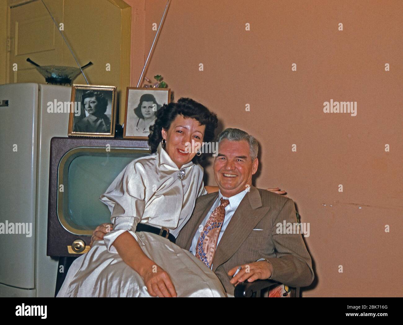An older happy couple in their home, USA, c 1955. The woman sits on the man's knee while he holds a cigarette. A TV set and a fridge are behind them in a room painted in colours typical of that era. This image is from an old American amateur Kodak colour transparency. Stock Photo