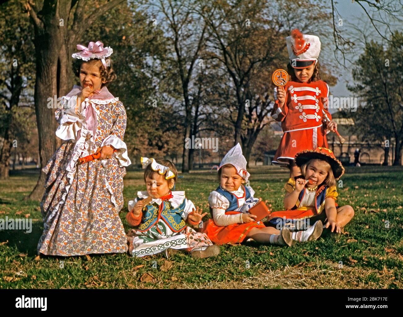 Five girls in a park in fancy dress costumes at Halloween, USA 1955. Three girls have orange popsicles (lollies) – two are licking them whilst the third (standing right in a marching-band uniform) holds hers up showing the image of a witch on it. Stock Photo