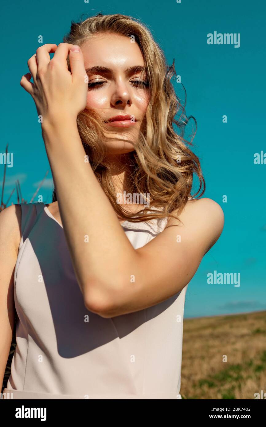 Portrait of a blonde young girl model who stands and poses on a sunny day against a background of haystacks and blue sky enjoying the rays of the sun. Stock Photo