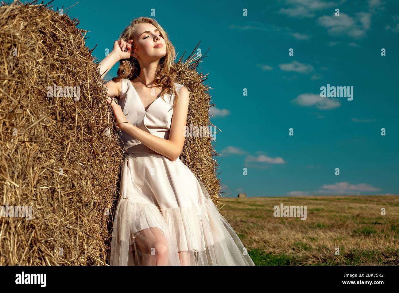 Portrait of a blonde young girl model who stands and poses on a sunny day against a background of haystacks and blue sky enjoying the rays of the sun. Stock Photo