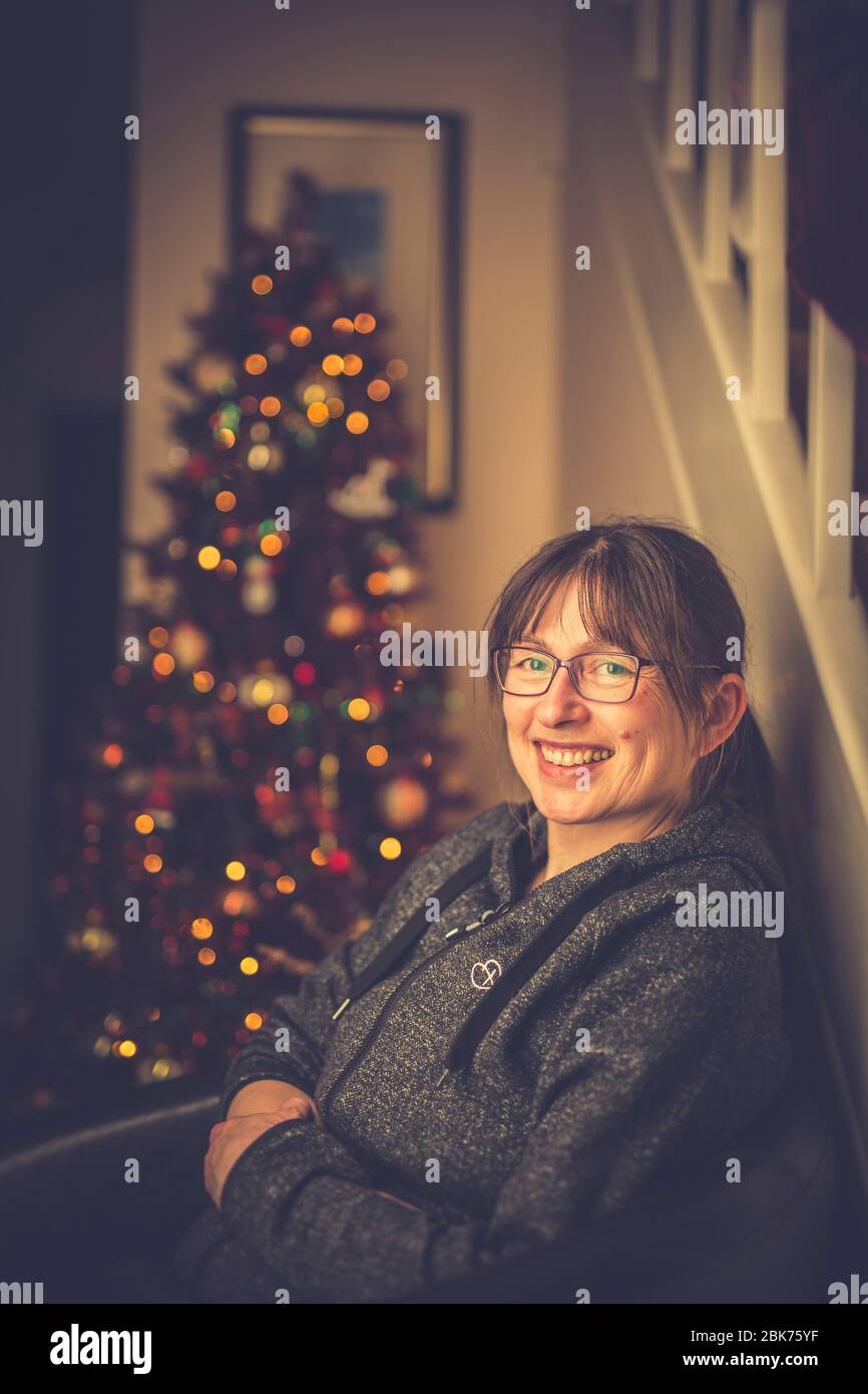 Close up of cheerful lady sitting in chair, relaxing in front of decorated Christmas tree looking pleased after cooking & eating the Christmas dinner. Stock Photo