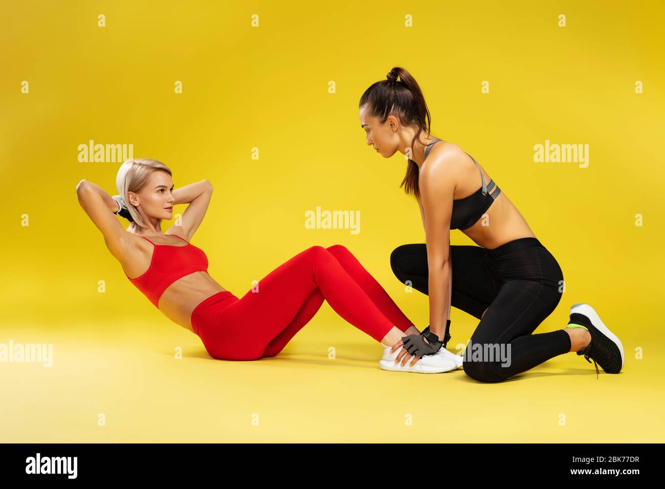 Fitness exercises. Attractive sporty blonde woman in red sportswear pumping press, exercising with personal trainer against yellow bakground Stock Photo