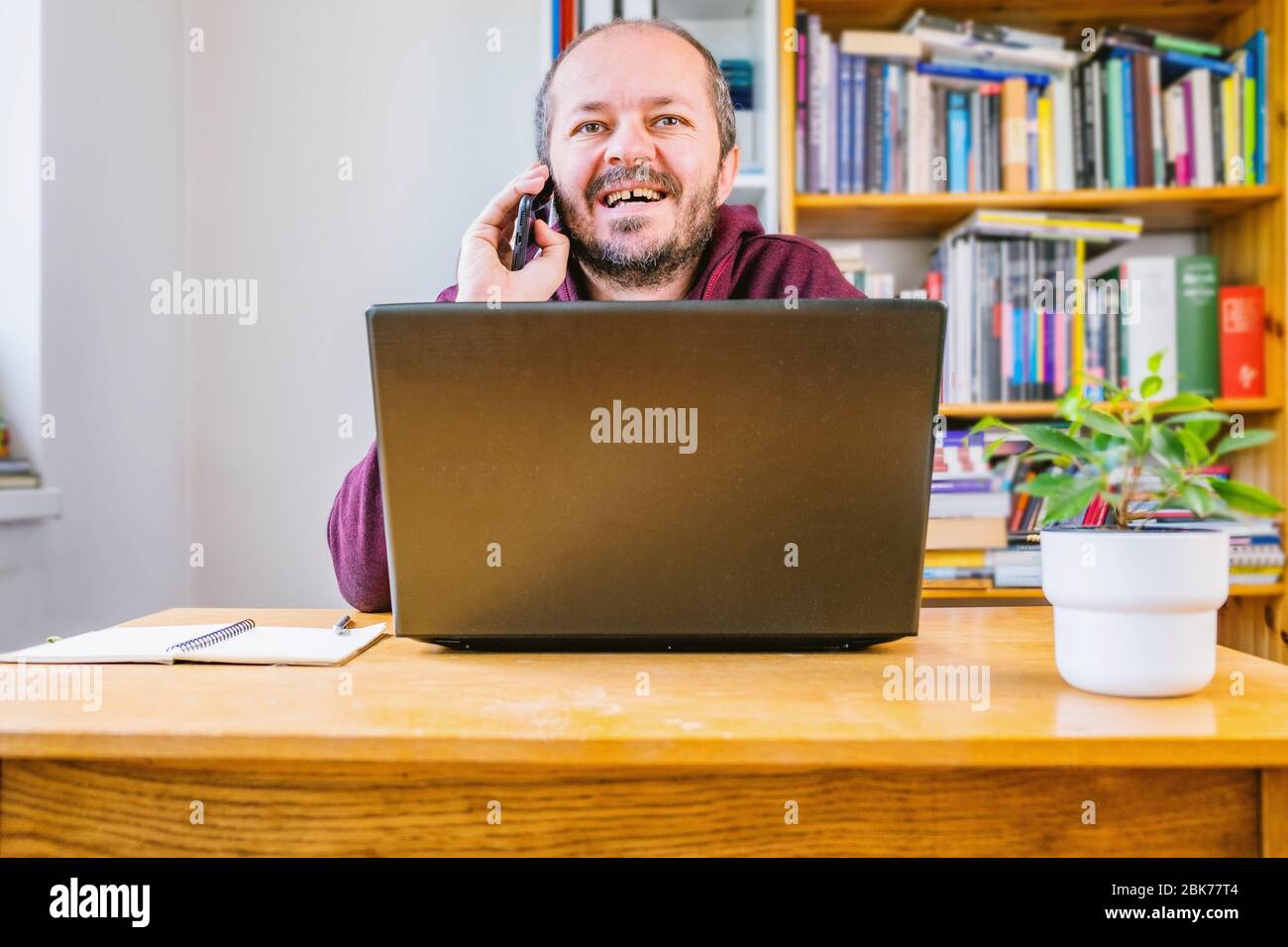 Home office. concept Bearded man sitting behind vintage desk at home, working on computer laptop and talking on smart phone, happy face expression Stock Photo