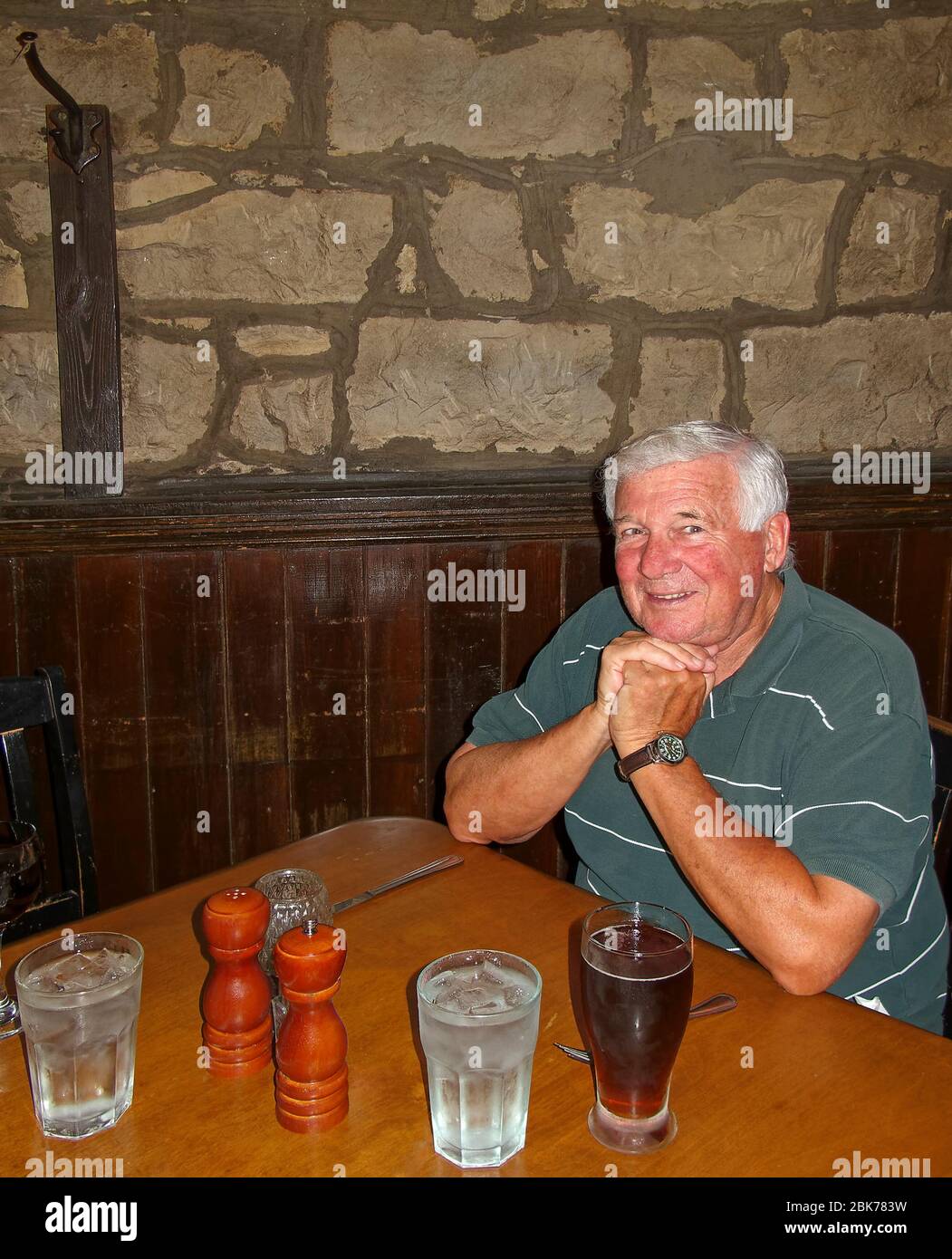 senior man, sitting at table, restaurant, hands under chin, smiling, happy, beer, water, large salt and pepper shakers, dark wood wainscoting, stone w Stock Photo