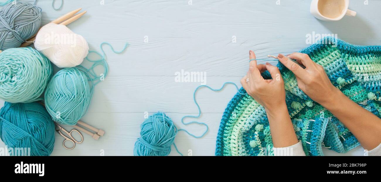 Women's hands are large. Woman crochets. Yarn of green, turquoise, aquamarine and blue colors. Blue wood long background. Stock Photo