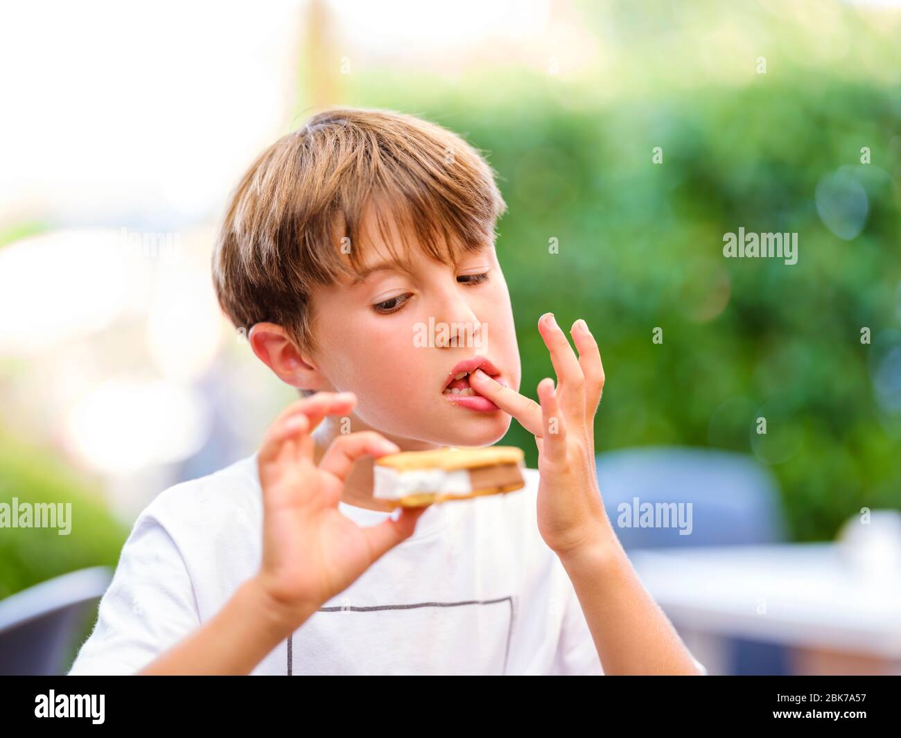 Close up portrait of beautiful boy licking finger while holding and eating sweet ice cream sandwich and looking at ice cream Stock Photo