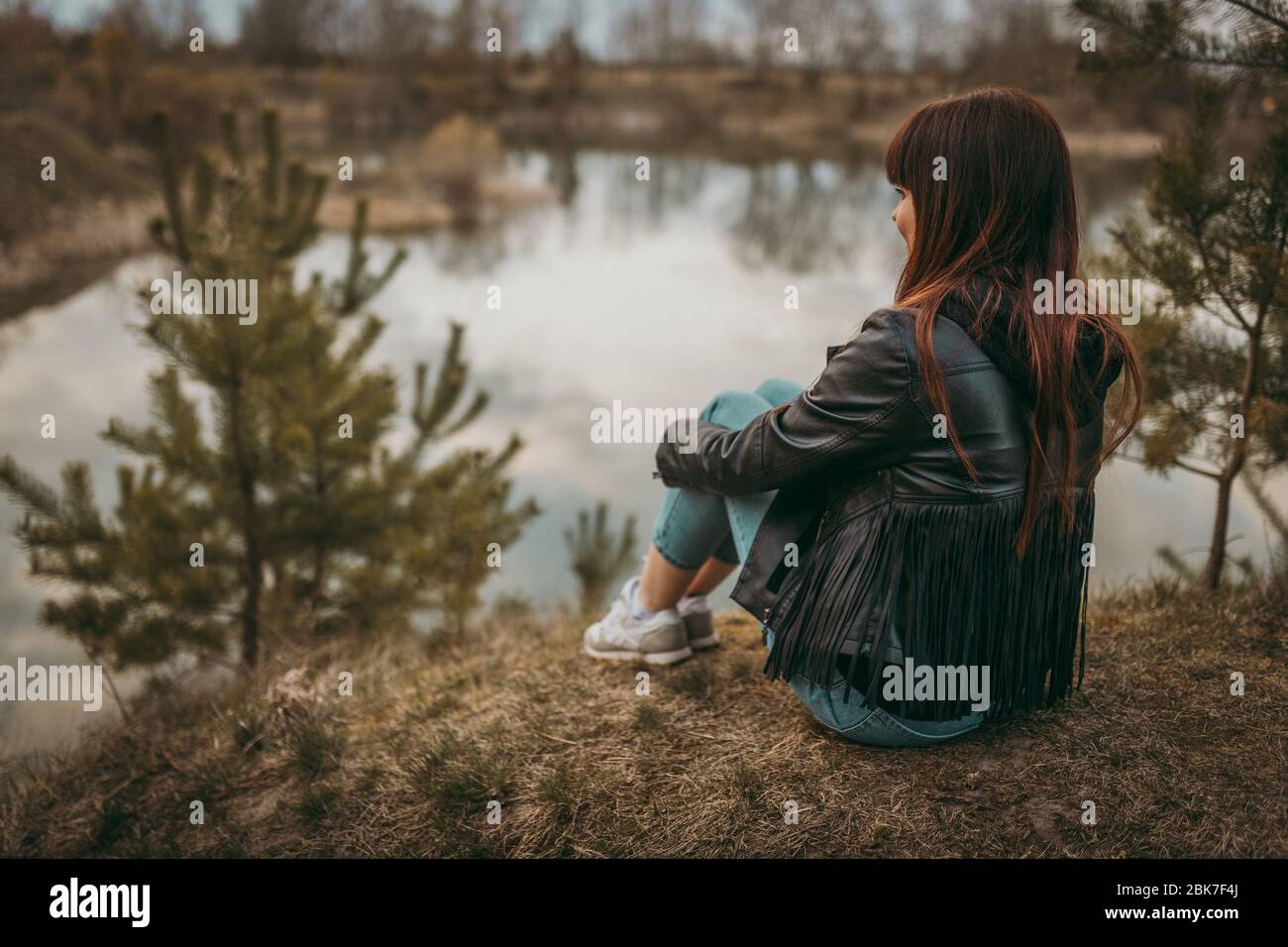 Beautiful sad girl sitting on the lake in jeans, looking into the ...