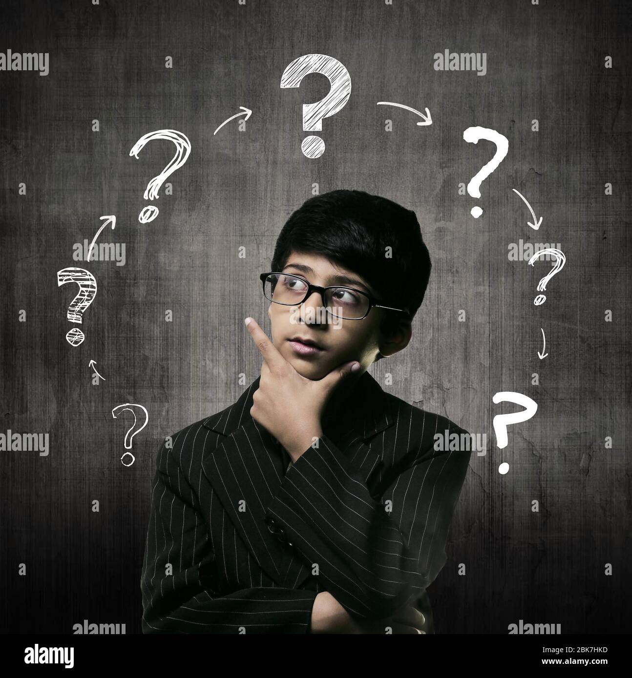 Cute Intelligent Little Boy Wearing Glasses, Hand Under Chin And Thinking While Standing Before A Chalkboard; Ring Of Question Marks Drawn On Chalk Bo Stock Photo