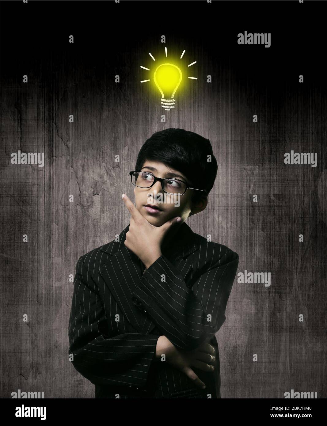 Cute Intelligent Little Boy Wearing Glasses, Hand Under Chin And Thinking While Standing Before A Chalkboard, Chalk Drawn Bulb Of Idea Stock Photo