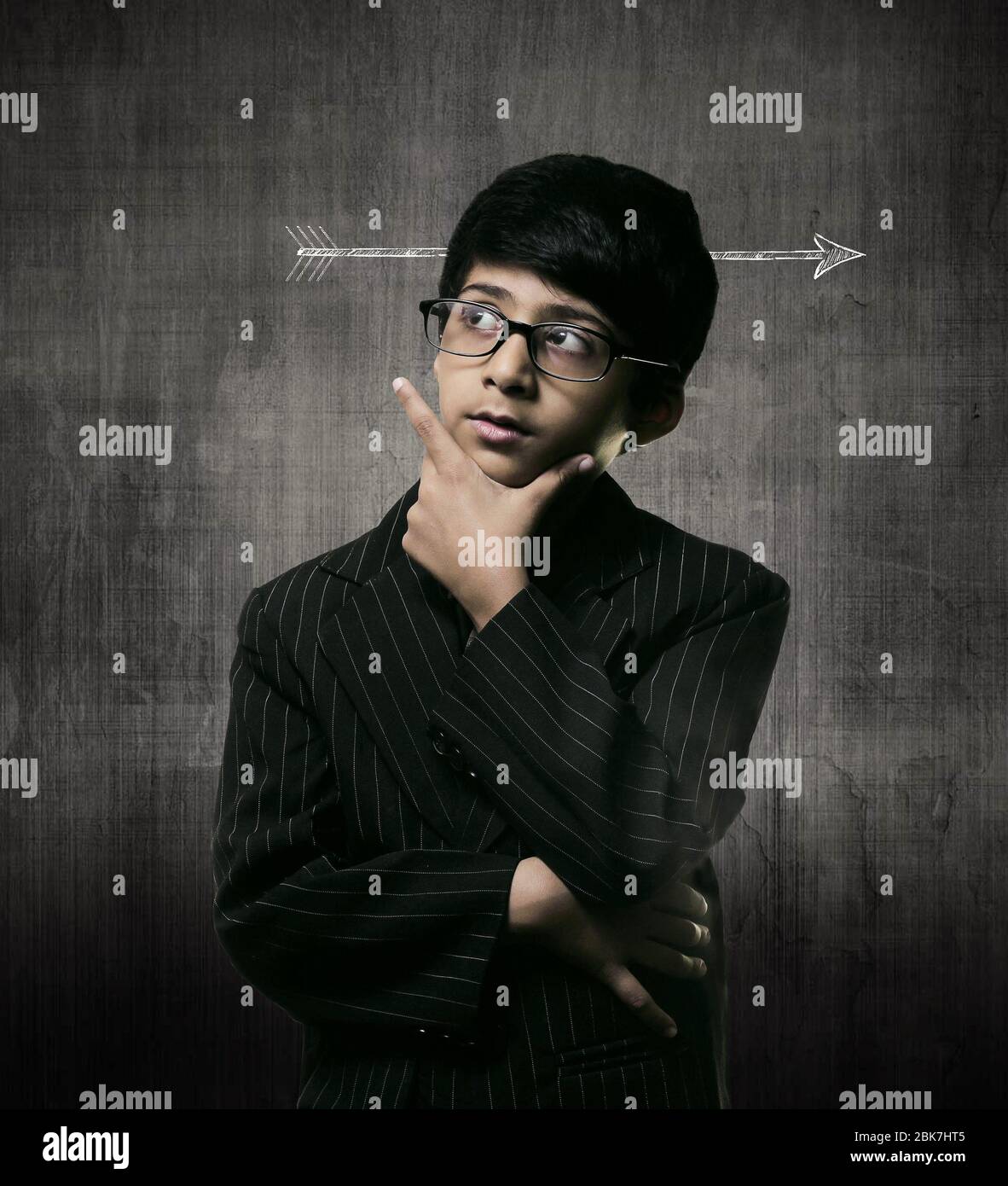 Cute Intelligent Little Boy Wearing Glasses, Hand Under Chin And Thinking While Standing Before A Chalkboard. Arrow Through Head Stock Photo